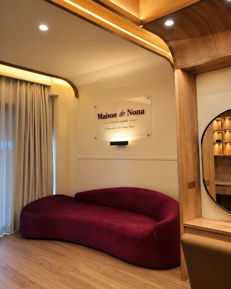 Area resepsionis: sofa beludru burgundy di bawah papan nama Maison de Nona, dengan pencahayaan hangat dan dinding berpanel kayu.